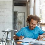 Hardworking excited African American A-student feeling happy after he solved complicated mathematical problem, smiling cheerfully and looking down in textbook while making notes in his copybook